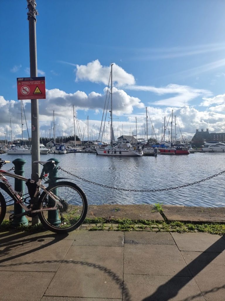 Large sailing boats glint in the sun at Ipswich Marina. A bike is parked up next to the water.