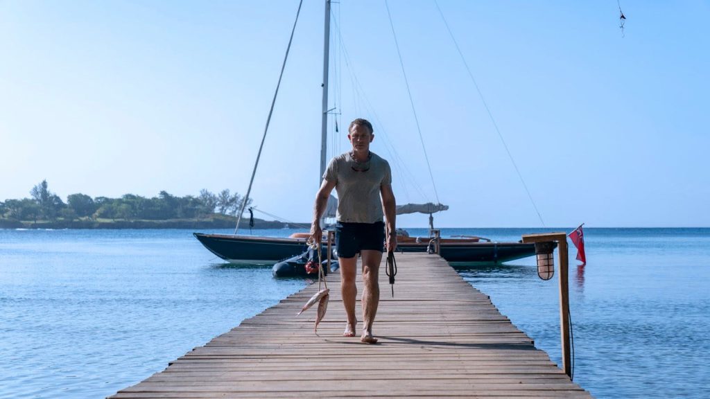 Daniel Craig as James Bond holds a bunch of fish and stands on a pier in front of a Sprit Yacht in the movie No Time to Die